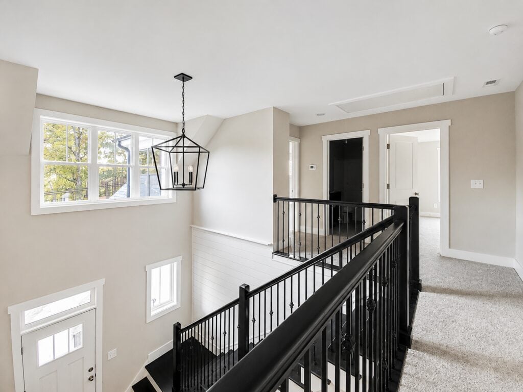 Expansive second-story loft space highlighting a black iron staircase railing, a large window, and a clear sightline toward the front entryway door below, crafted by Travars Built Homes.