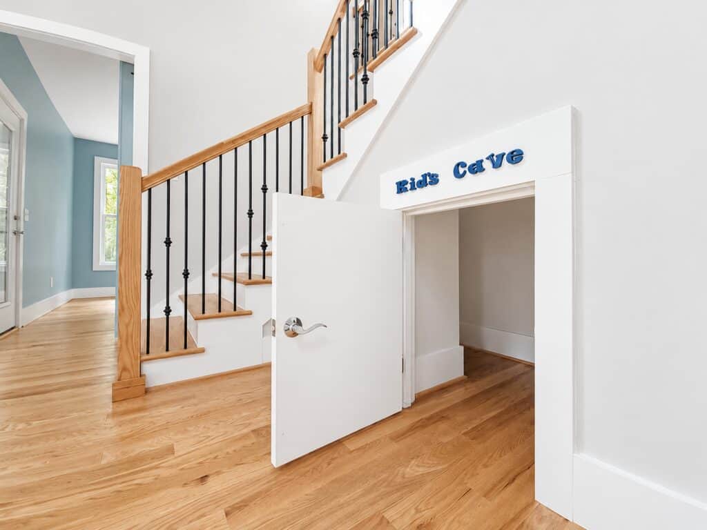 Delightful custom playroom entry built directly under a wood-and-iron staircase, featuring a miniature white door labeled "Kid's Cave" opening onto oak hardwood floors, by Travars Built Homes.