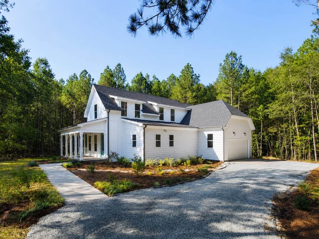 Beautiful side-angle view of a custom modern farmhouse surrounded by mature woods, featuring white horizontal and vertical siding, a deep wraparound porch, and a side-load garage, by Travars Built Homes.