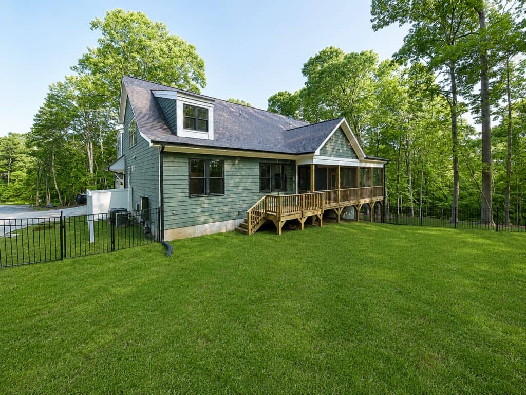 Custom home exterior featuring sage green horizontal lap siding, striking black window frames, and a wooden staircase leading up to a covered screened porch, surrounded by a fenced wooded lot, designed by Travars Built Homes.
