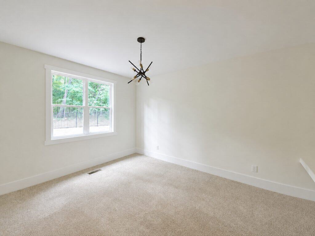Minimalist custom bedroom highlighting soft neutral carpeting, a black geometric chandelier, and a bright window offering views of the wooded backyard, designed by Travars Built Homes.