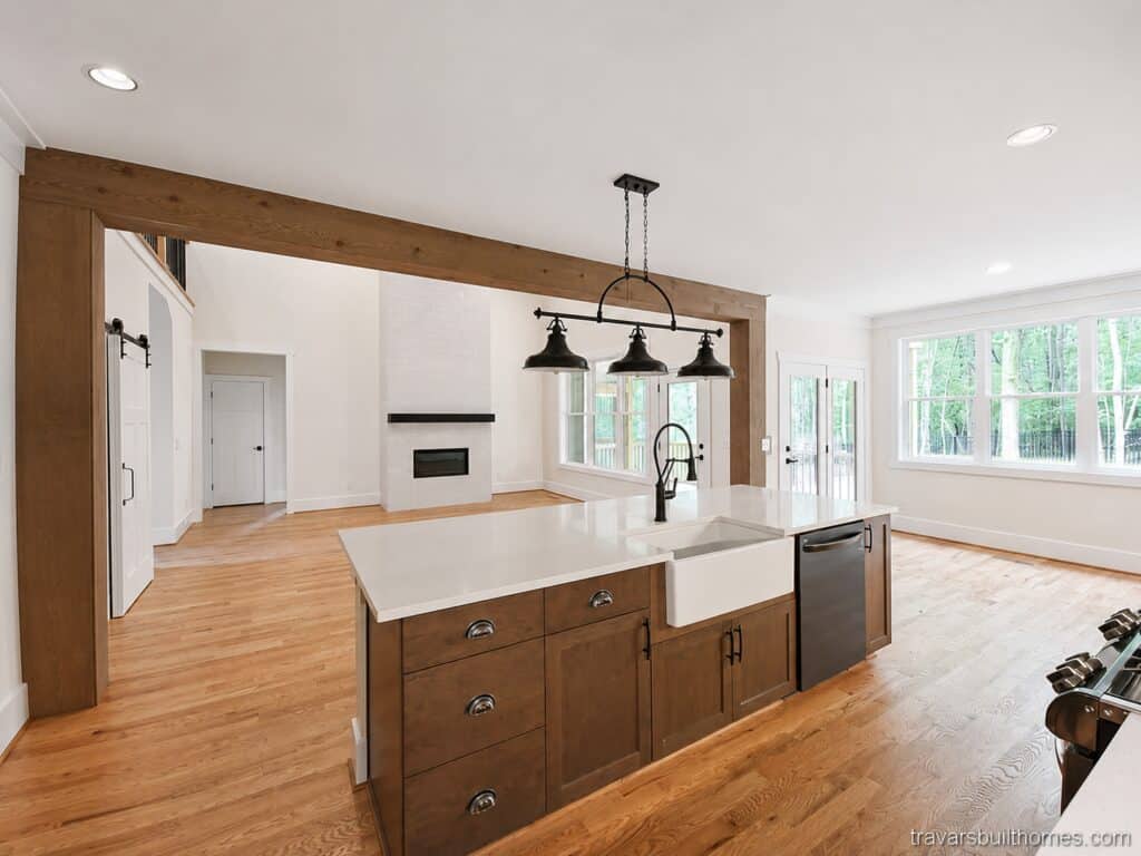 Custom kitchen design highlighting a dark wood island with a white farmhouse sink, a classic herringbone backsplash, and a clear sightline through a rustic wood beam into a two-story living room, built by Travars Built Homes.