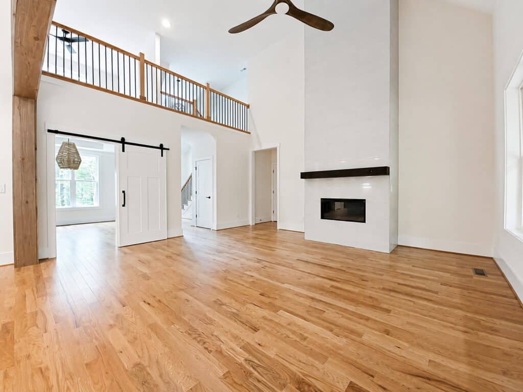 Expansive view of a custom two-story living room featuring rich oak hardwood floors, a towering white tile fireplace surround, and an overlooking loft with wood and iron railings, built by Travars Built Homes.