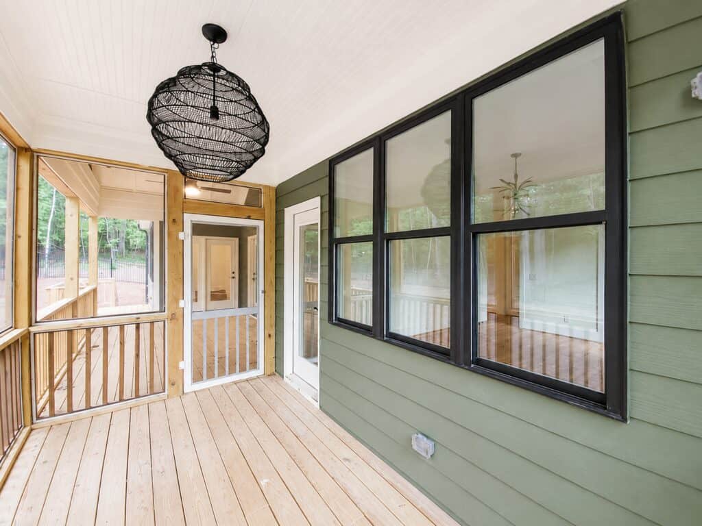 Detail view of a custom screened-in porch featuring a sage green exterior wall, large black-framed windows looking into the home, a white beadboard ceiling, and a woven pendant light, designed by Travars Built Homes.