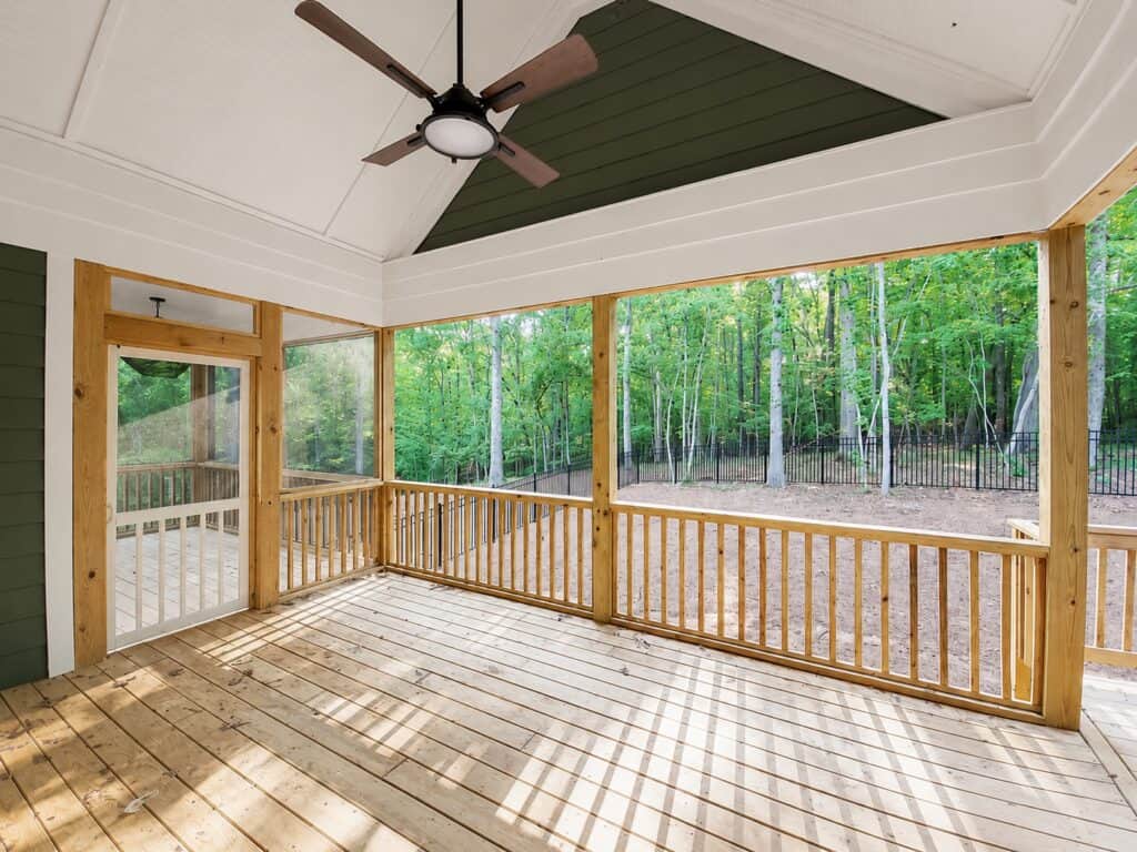 Wide view of a custom screened-in porch showcasing natural wood decking, a dramatic dark green vaulted ceiling with a modern black fan, and expansive screens framing a lush wooded backyard, by Travars Built Homes.