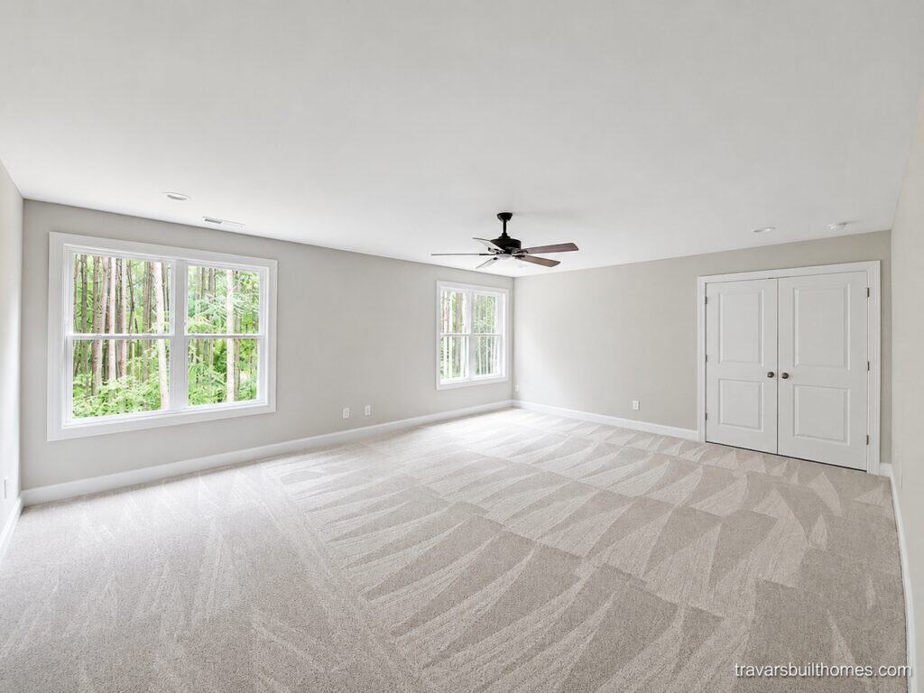 Expansive finished bonus room highlighting plush patterned carpeting, soft light grey walls, multiple windows viewing the tree canopy, a modern ceiling fan, and double doors for ample storage space, built by Travars Built Homes.