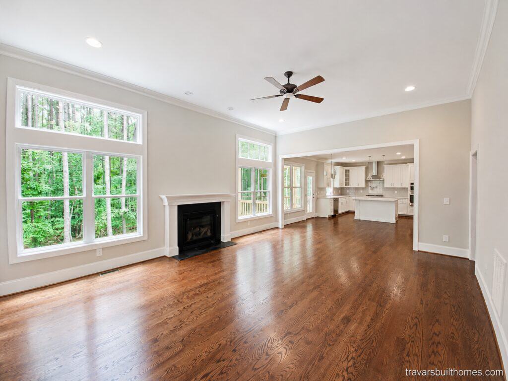 Expansive open-concept living area featuring rich hardwood floors, a classic white fireplace mantel, large picture windows framing woodland views, and a seamless flow into a bright custom kitchen, built by Travars Built Homes.