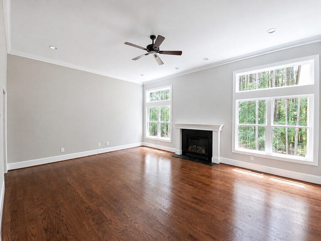 Bright and airy custom living room showcasing a classic white fireplace, rich oak hardwood floors, a modern black ceiling fan, and large picture windows framing a wooded backyard, by Travars Built Homes.