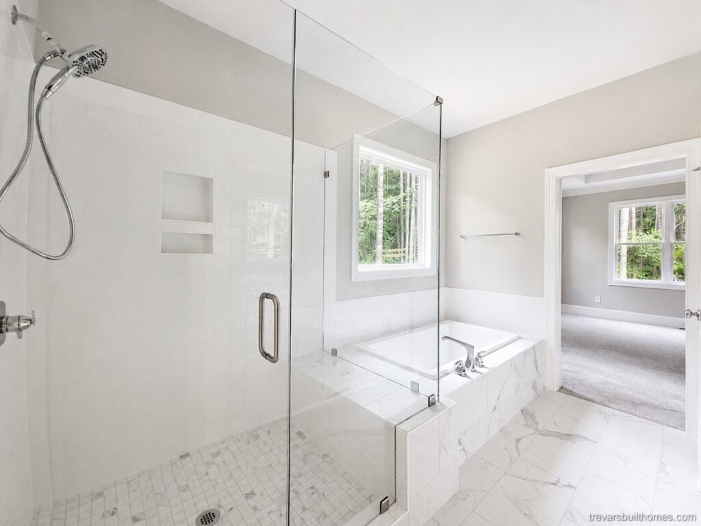 Luxurious custom bathroom highlighting a seamless glass walk-in shower connected to a tiled drop-in soaking tub beneath a bright window, designed by Travars Built Homes.