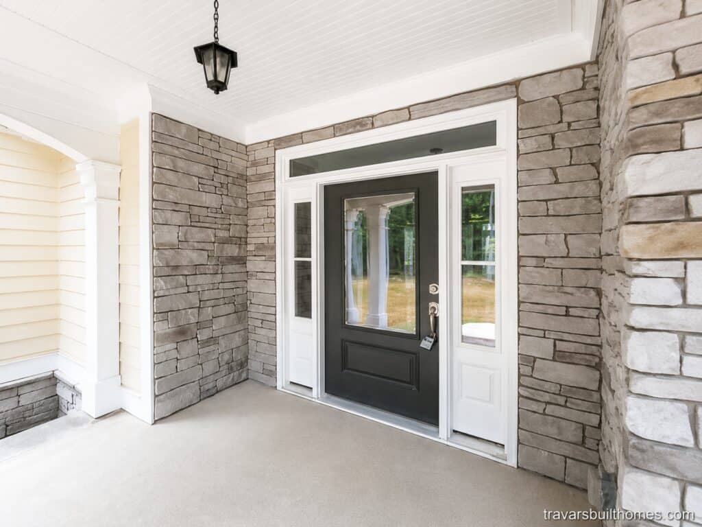 Elegant custom front entry porch highlighting a black glass-paneled front door with matching sidelights, set within a beautiful grey stacked stone exterior facade, designed by Travars Built Homes.