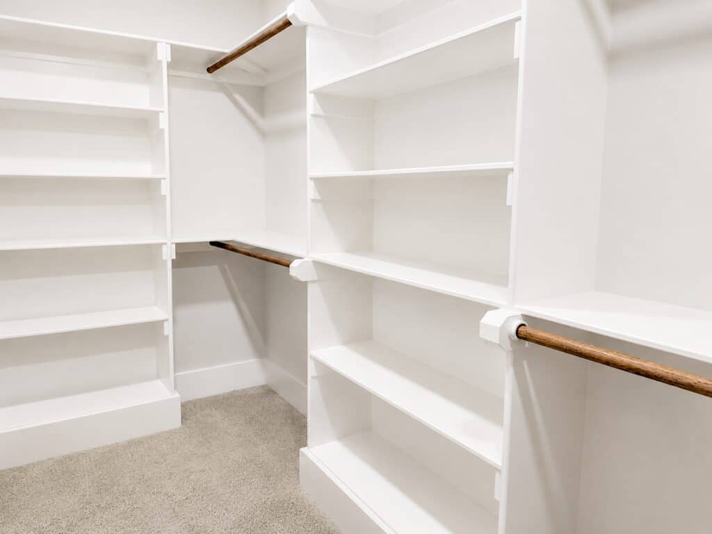 Custom primary walk-in closet detailing extensive built-in white laminate shelving, wooden hanging rods, and soft grey carpeting, crafted by Travars Built Homes.