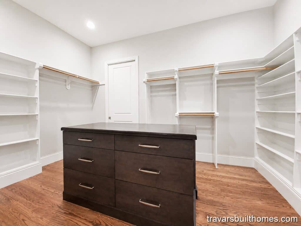Spacious custom master walk-in closet showcasing extensive white built-in shelving and wooden hanging rods surrounding a dark wood center island dresser, resting on oak hardwood floors, built by Travars Built Homes.