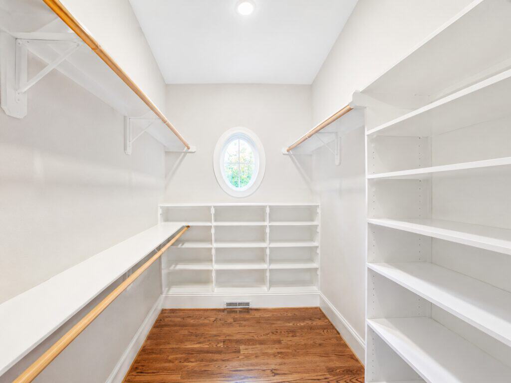 Custom master walk-in closet design featuring continuous white built-in shelving, wooden hanging rods, rich oak hardwood floors, and a unique oval accent window, built by Travars Built Homes.