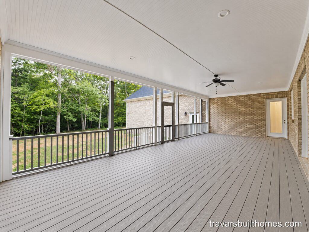 Large custom screened-in back porch highlighting grey decking, a white beadboard ceiling with a black ceiling fan, and a light brick exterior wall, built by Travars Built Homes.