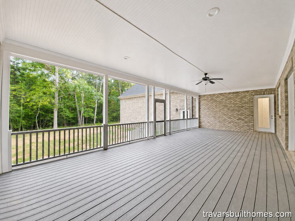 Wide view of a spacious custom screened porch featuring light grey decking, a white beadboard ceiling with a modern fan, and expansive screens framing a lush wooded backyard, by Travars Built Homes.