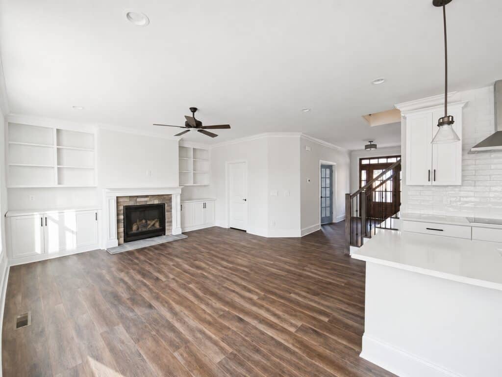 Expansive view of a custom open-concept living area, highlighting a stone fireplace with white built-ins, rich wood-look flooring, and a clear sightline to the front entry and open staircase, built by Travars Built Homes.