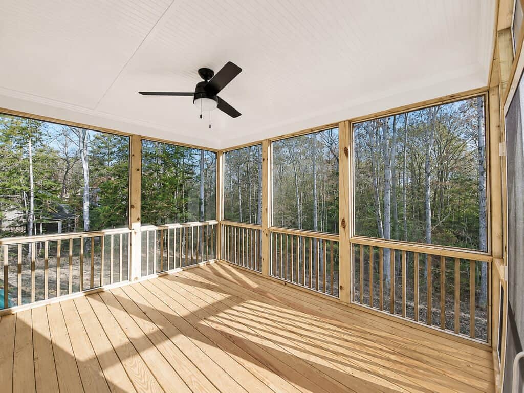 Custom screened outdoor living space featuring natural pine decking, a white beadboard ceiling with a modern black fan, and panoramic views of the surrounding woodland, built by Travars Built Homes.
