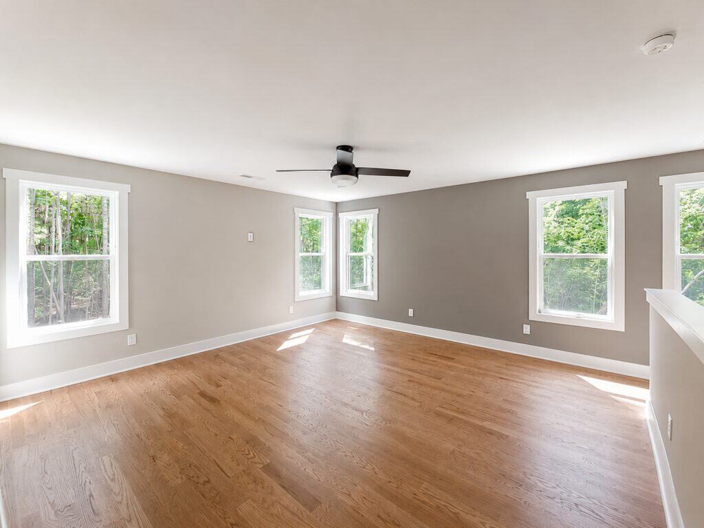Bright finished bonus room featuring warm oak hardwood floors, soft greige walls, multiple windows providing natural light, and a modern dark ceiling fan, crafted by Travars Built Homes.