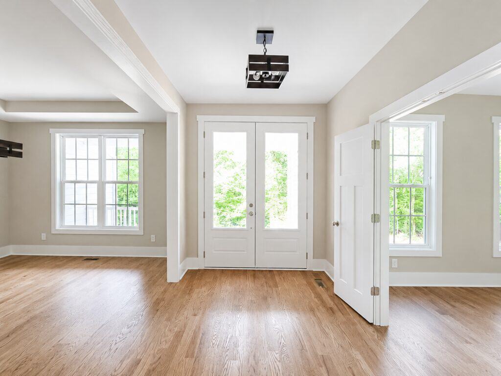 Open and airy custom front entryway highlighting solid oak hardwood floors, elegant white double front doors with large glass panes, and a modern dark metal square chandelier, designed by Travars Built Homes.