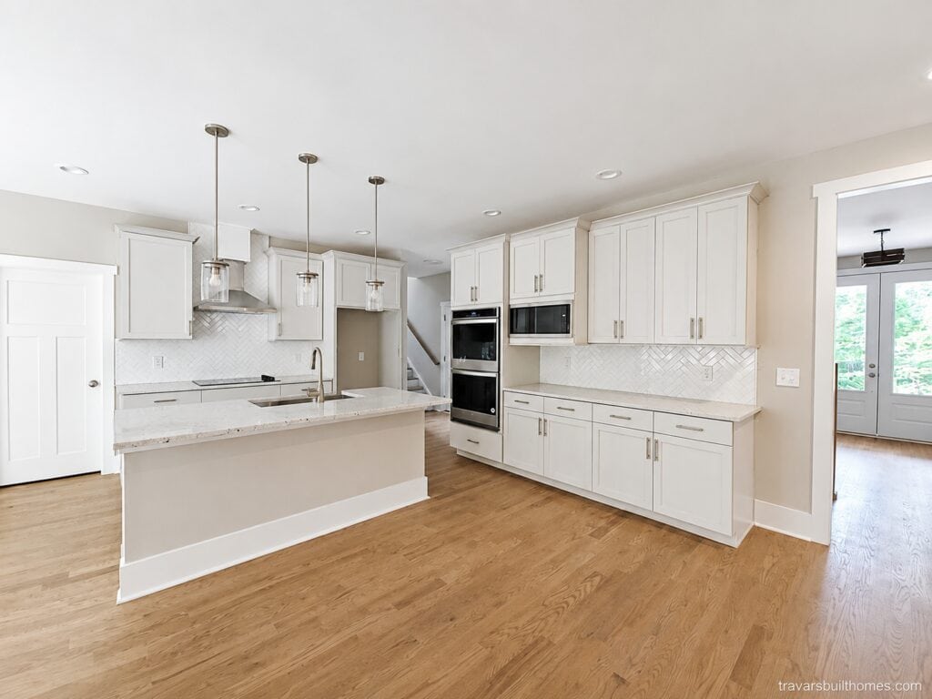 Custom kitchen workspace featuring pristine white shaker cabinetry, a large granite-topped island, a white herringbone tile backsplash, and built-in stainless steel appliances, crafted by Travars Built Homes.