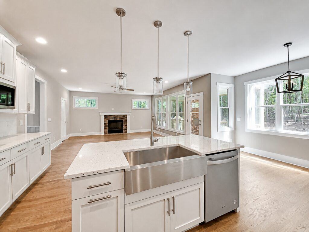 Bright custom kitchen space featuring crisp white shaker cabinetry, a large island with a stainless steel farmhouse sink and quartz countertops, clear glass pendants, and a view into the family room, built by Travars Built Homes.