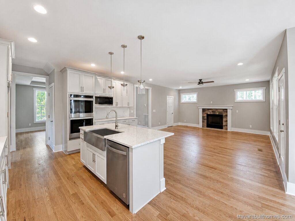 Expansive view from a custom kitchen island with an apron-front stainless steel sink, looking across rich oak floors into an open living room centered around a brick fireplace, built by Travars Built Homes.