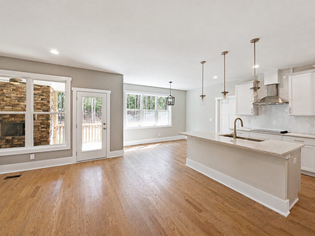 Open-concept kitchen and dining area showcasing a white island with granite countertops, sleek pendant lights, and a clear view into a cozy keeping room with a stacked stone fireplace, designed by Travars Built Homes.