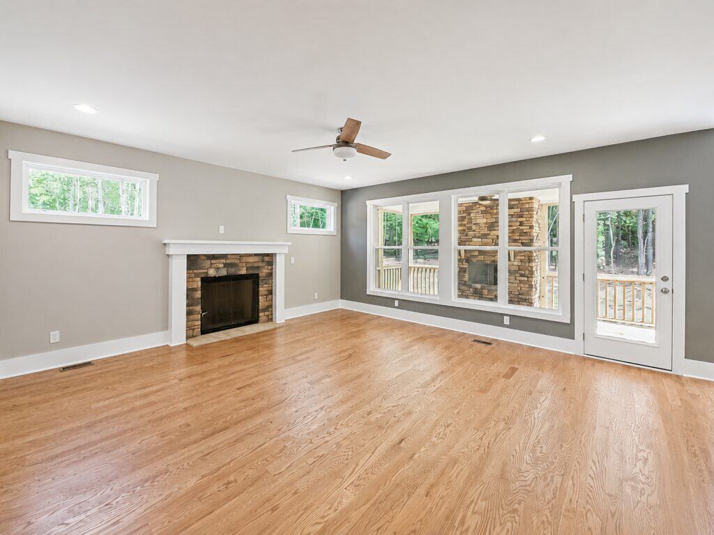 Bright custom living room highlighting warm oak hardwood floors, a classic brick fireplace with a crisp white mantel, large rear-facing windows, and a glass door leading to an outdoor living space, by Travars Built Homes.