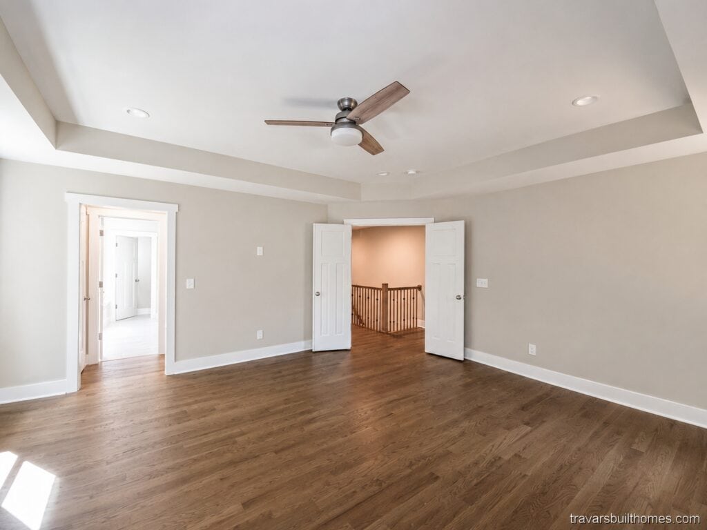 Spacious custom master bedroom featuring rich hardwood floors, soft greige walls, an elegant tray ceiling with a modern fan, and wide double doors opening into a hallway, built by Travars Built Homes.