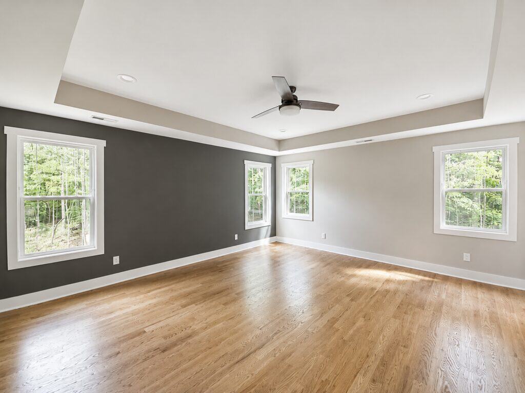 Custom primary bedroom showcasing a bold charcoal accent wall, contrasting light grey walls, a tray ceiling with a ceiling fan, and beautiful natural oak hardwood floors, built by Travars Built Homes.
