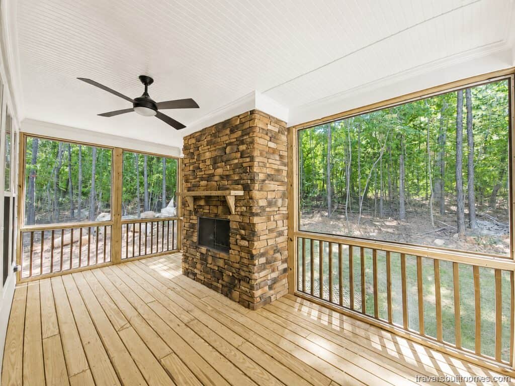 Cozy screened-in porch showcasing a floor-to-ceiling stacked stone fireplace, natural timber flooring, a white beadboard ceiling, and a modern dark fan, surrounded by forest views, built by Travars Built Homes.