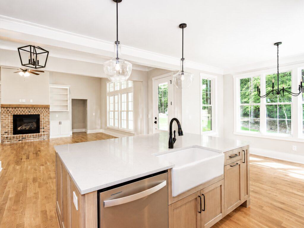 Bright, open-concept custom kitchen highlighting a light wood island with an apron-front farmhouse sink, clear glass pendant lighting, and a clear view into a living area with a brick fireplace, by Travars Built Homes.