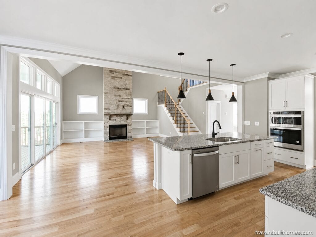 Custom open-concept view looking across a granite-topped kitchen island with dark pendant lights toward a bright living room centered around a stacked stone fireplace, built by Travars Built Homes.