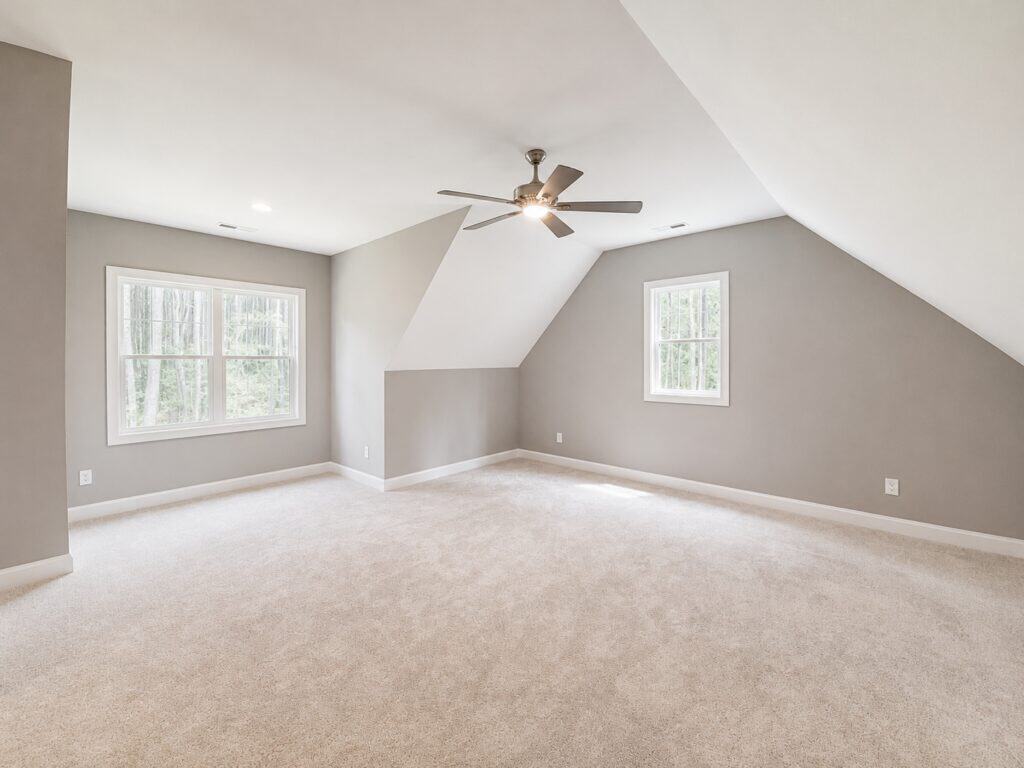 Finished custom bonus room featuring soft beige-grey walls, plush beige carpeting, angled dormer ceilings, a classic ceiling fan, and multi-pane windows, by Travars Built Homes.