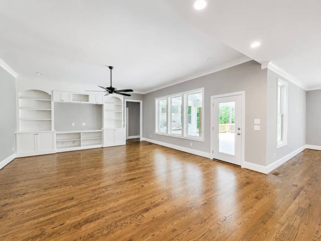 Spacious open great room configuration showcasing natural oak floors, a large white custom media bookcase unit, and an array of windows viewing out to a back porch, designed by Travars Built Homes.
