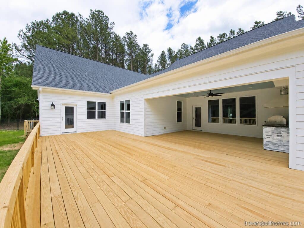 Expansive custom wood deck wrapping around the rear of a white farmhouse, highlighting a large covered patio section equipped with an outdoor kitchen setup, built by Travars Built Homes.