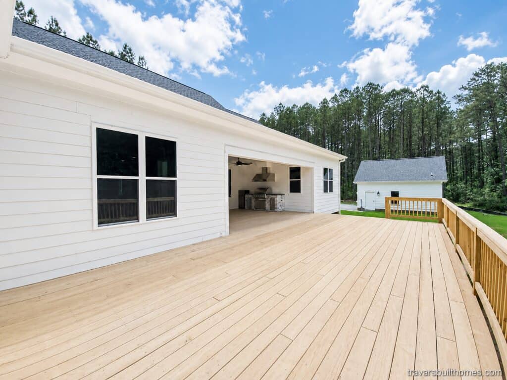 View along an extensive open-air timber deck, highlighting a deep covered porch alcove with a built-in outdoor kitchen against a crisp white farmhouse exterior, by Travars Built Homes.