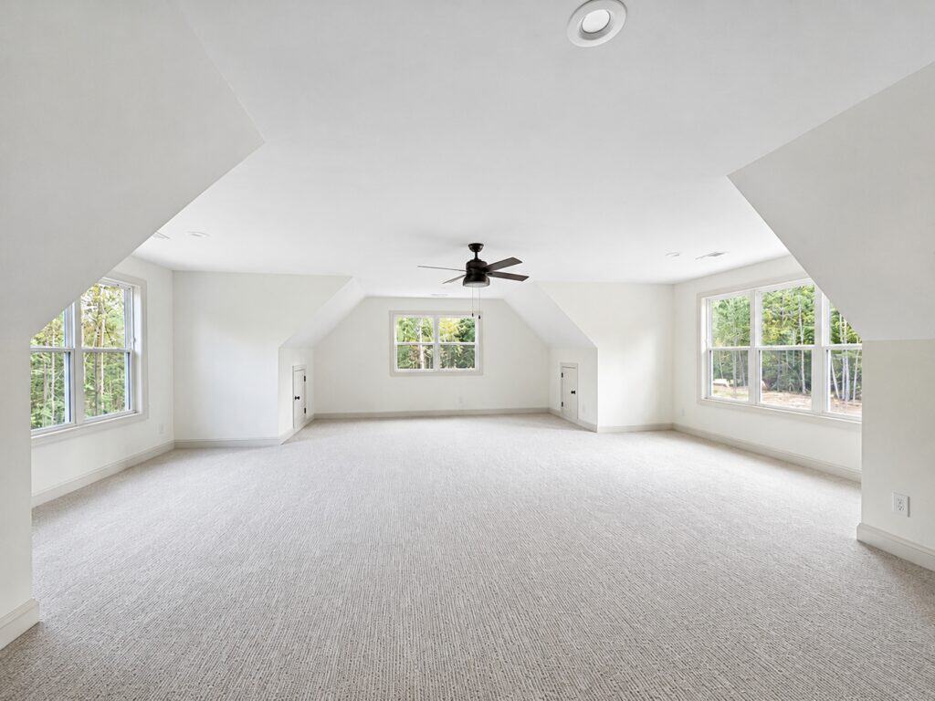 Generous finished custom bonus room featuring soft gray carpeting, angled dormer ceilings, built-in storage access, and a black ceiling fan, built by Travars Built Homes.