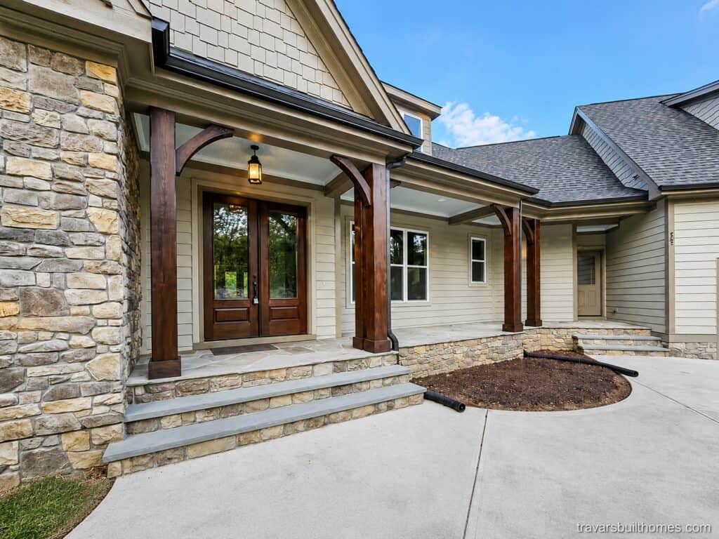 Close-up of a custom front entry featuring a stone veneer facade, a covered porch supported by heavy timber columns with brackets, and a dark wood double front door, built by Travars Built Homes.