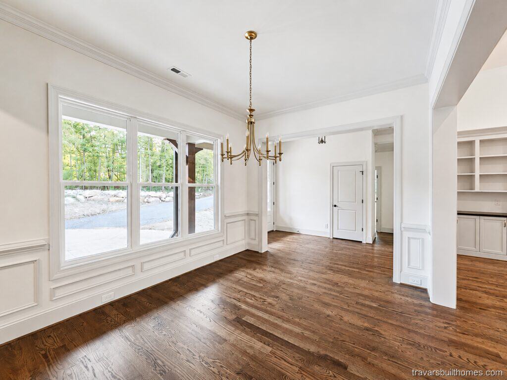 Bright custom formal dining space featuring a classic metal chandelier, rich oak floors, formal wainscoting trim, and large windows that fill the open room with natural light, by Travars Built Homes.
