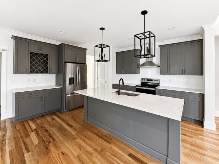 Custom kitchen design featuring sophisticated dark gray shaker cabinets, a built-in wine rack, a matching center island with a white sink, and stylish geometric black pendant lights, crafted by Travars Built Homes.