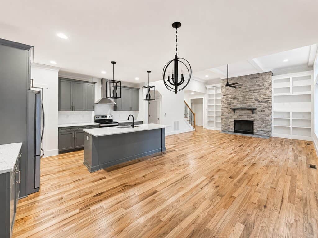 Expansive custom open floor plan viewing from a modern kitchen with gray cabinets and a central island into a spacious living room centered around a stone fireplace, by Travars Built Homes.