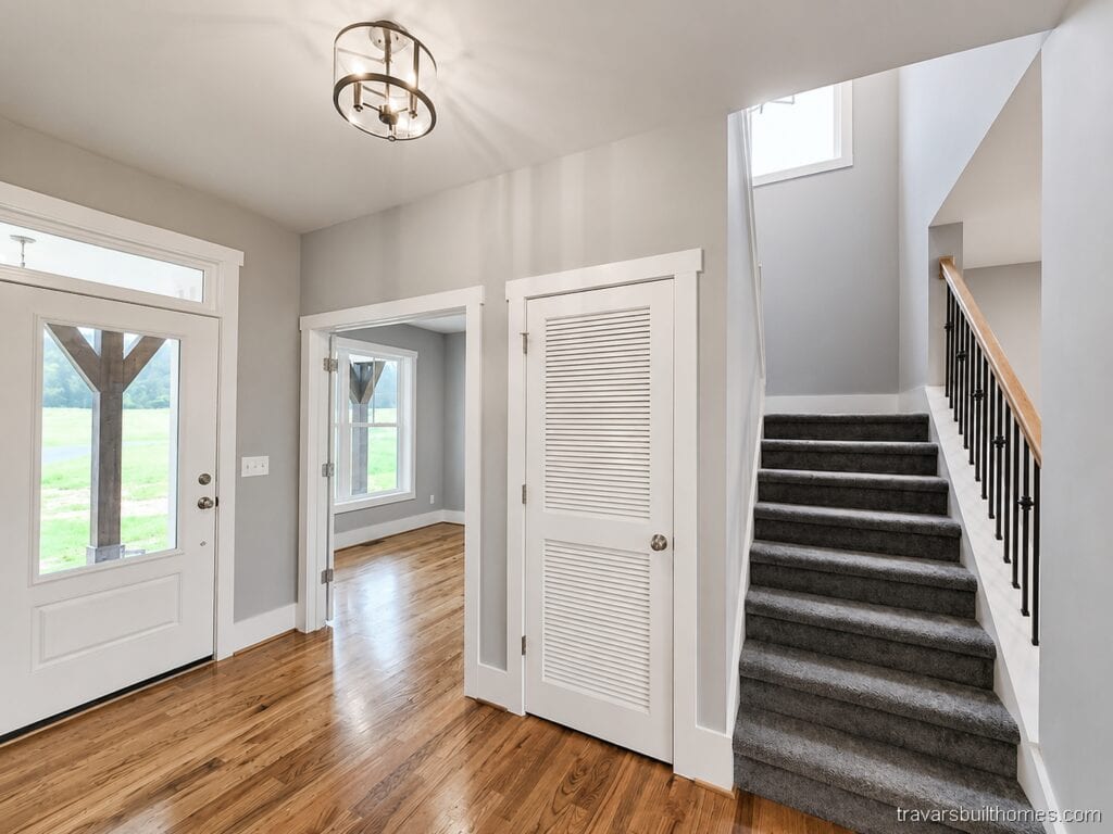Custom home front entry foyer showcasing a white front door with sidelights, warm hardwood flooring, grey walls, and a carpeted staircase with dark iron balusters, by Travars Built Homes.