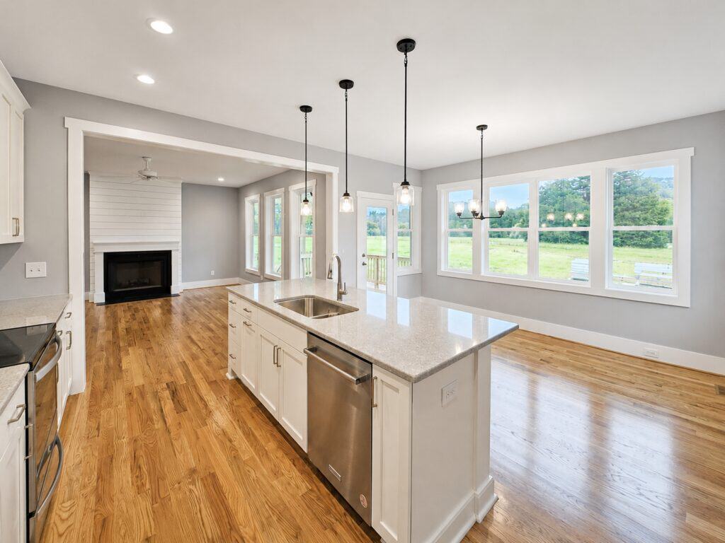 Bright open-concept custom kitchen highlighting a central island with white quartz countertops, sleek pendant lights, a large dining area with field views, and an open layout leading into a fireplace living room, by Travars Built Homes.