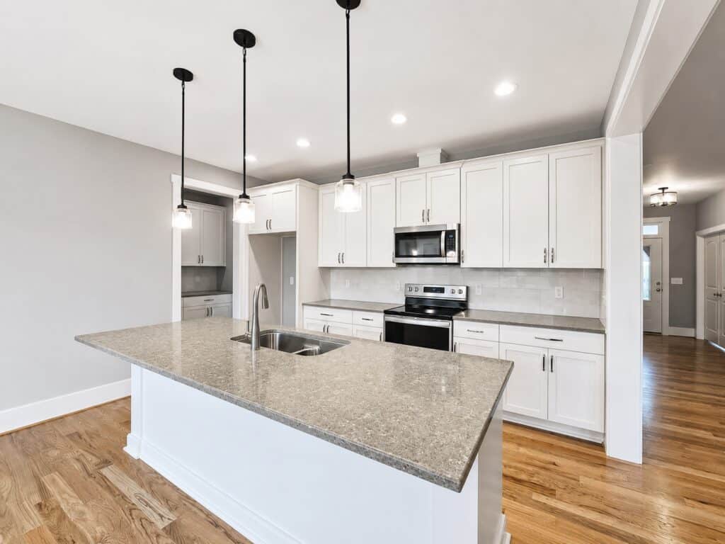 Custom kitchen design featuring white shaker cabinets, gray quartz countertops, stainless steel appliances, a tile backsplash, and a large center island with black glass pendant lights, built by Travars Built Homes.