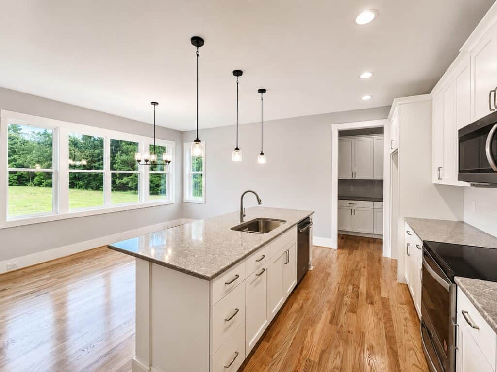Spacious open kitchen looking toward a scenic breakfast nook framed by large windows, with a view of a walk-in scullery pantry from behind the quartz-topped central island, crafted by Travars Built Homes.