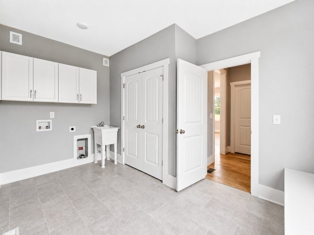 Custom laundry room showcasing gray tile floors, light gray walls, white storage cabinets, a standalone utility sink, and dual white six-panel closet doors, designed by Travars Built Homes.