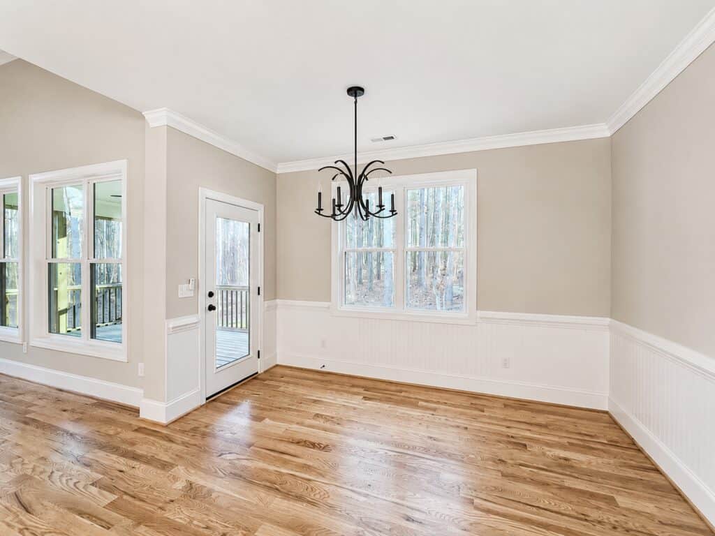 Bright custom breakfast nook highlighting warm hardwood floors, white beadboard wainscoting, an elegant black chandelier, and a glass-paneled door leading to the porch, designed by Travars Built Homes.