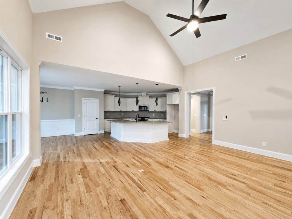 Open concept custom living room featuring a vaulted ceiling with a modern fan, warm hardwood floors, and a seamless flow into a bright kitchen with a large island, by Travars Built Homes.
