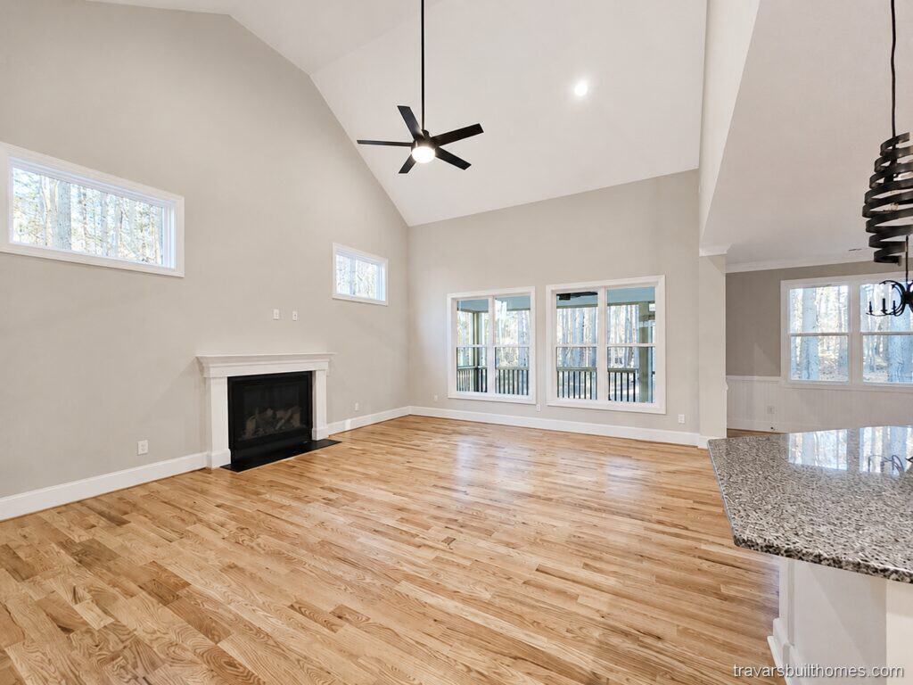 Spacious custom living room highlighting warm hardwood floors, a vaulted ceiling with a modern black fan, and a fireplace with a white mantel and black granite surround, by Travars Built Homes.